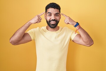Hispanic man with beard standing over yellow background smiling pointing to head with both hands finger, great idea or thought, good memory