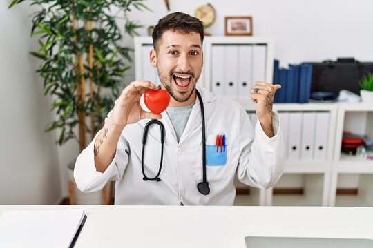 Young doctor holding heart at medical clinic pointing thumb up to the side smiling happy with open mouth