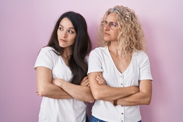 Mother and daughter standing together over pink background looking to the side with arms crossed convinced and confident
