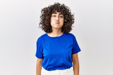 Young middle eastern woman standing over isolated background puffing cheeks with funny face. mouth inflated with air, crazy expression. - obrazy, fototapety, plakaty