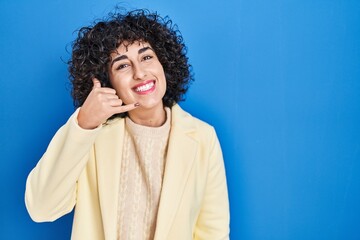 Fototapeta na wymiar Young brunette woman with curly hair standing over blue background smiling doing phone gesture with hand and fingers like talking on the telephone. communicating concepts.
