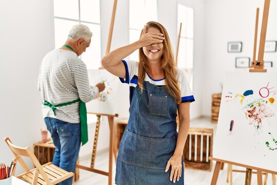 Hispanic woman wearing apron at art studio smiling and laughing with hand on face covering eyes for surprise. blind concept.