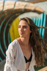 Chica joven con pelo largo sonriendo y posando con cascos de musica delante de tablas de surf en la playa