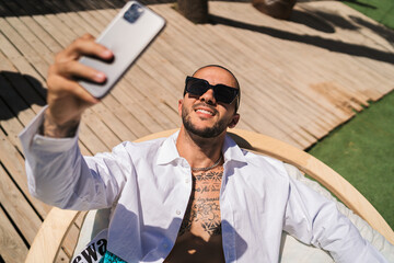 Chico joven musculoso con la camisa abierta haciéndose selfie en una silla de un bar de playa