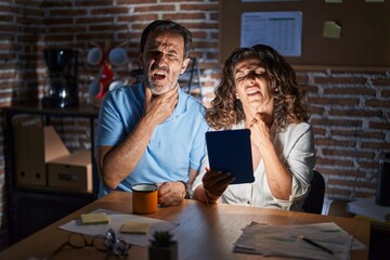 Middle age hispanic couple using touchpad sitting on the table at night touching painful neck, sore throat for flu, clod and infection