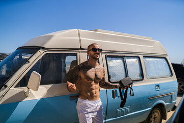 Chico joven tatuado y musculoso posando delante de una furgoneta celeste y blanca en zona de campers