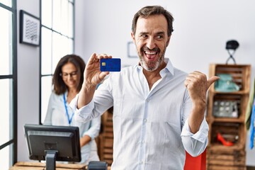Hispanic man holding credit card at retail shop pointing thumb up to the side smiling happy with open mouth