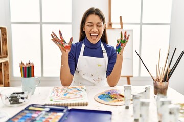 Young brunette woman at art studio with painted hands celebrating mad and crazy for success with...