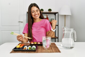 Obraz na płótnie Canvas Young brunette woman eating sushi using chopsticks very happy and excited doing winner gesture with arms raised, smiling and screaming for success. celebration concept.