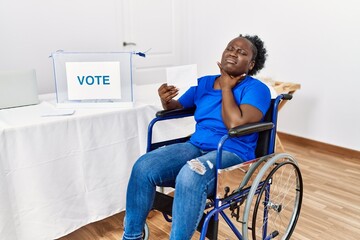 Young african woman sitting on wheelchair voting putting envelop in ballot box touching painful...