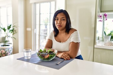 Obraz na płótnie Canvas Young hispanic woman eating healthy salad at home relaxed with serious expression on face. simple and natural looking at the camera.