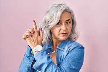 Middle age woman with grey hair standing over pink background holding symbolic gun with hand gesture, playing killing shooting weapons, angry face - Powered by Adobe