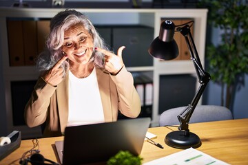 Middle age woman with grey hair working using computer laptop late at night smiling cheerful showing and pointing with fingers teeth and mouth. dental health concept.