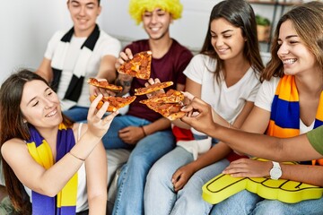 Group of young friends watching and supporting soccer match eating pizza at home.