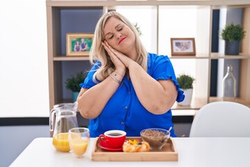 Obraz na płótnie Canvas Caucasian plus size woman eating breakfast at home sleeping tired dreaming and posing with hands together while smiling with closed eyes.