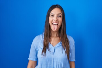Fototapeta na wymiar Young brunette woman standing over blue background sticking tongue out happy with funny expression. emotion concept.