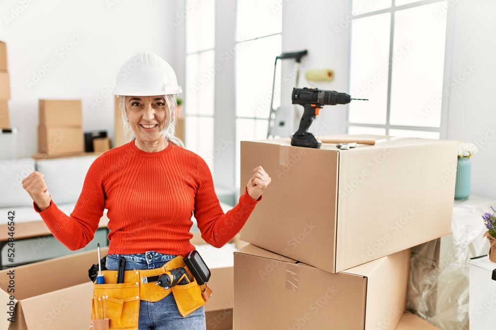 Poster middle age grey-haired woman wearing hardhat standing at new home very happy and excited doing winne