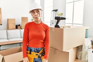 Middle age grey-haired woman wearing hardhat standing at new home making fish face with lips, crazy and comical gesture. funny expression.