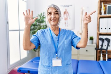 Middle age grey-haired woman wearing physiotherapist uniform at medical clinic showing and pointing up with fingers number seven while smiling confident and happy.