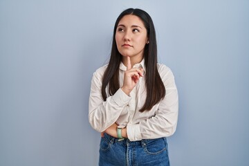 Young latin woman standing over blue background thinking concentrated about doubt with finger on...