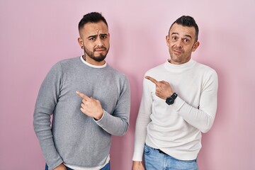 Homosexual couple standing over pink background pointing aside worried and nervous with forefinger,...