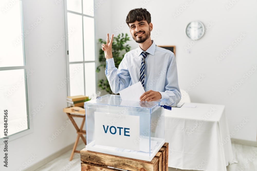 Sticker Hispanic man with beard voting putting envelop in ballot box smiling looking to the camera showing fingers doing victory sign. number two.