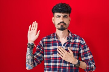 Young hispanic man with beard standing over red background swearing with hand on chest and open palm, making a loyalty promise oath