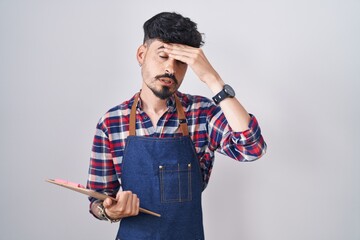Young hispanic man with beard wearing waiter apron holding clipboard worried and stressed about a...