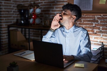 Young hispanic man with beard working at the office at night shouting and screaming loud to side with hand on mouth. communication concept.