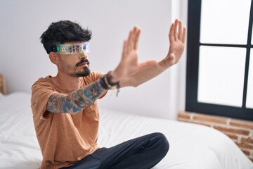 Young hispanic man using augmented reality glasses sitting on bed at bedroom