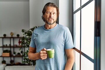 Middle age caucasian man drinking coffee leaning on the window at home