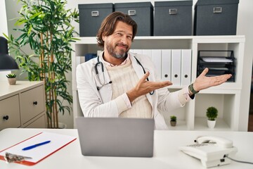 Handsome middle age doctor man working at the clinic inviting to enter smiling natural with open hand