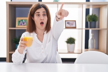 Brunette woman drinking glass of orange juice pointing with finger surprised ahead, open mouth amazed expression, something on the front