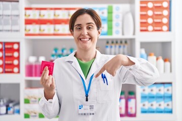 Brunette woman working at pharmacy drugstore holding condom pointing finger to one self smiling...