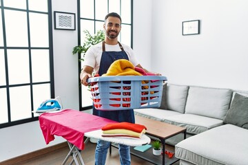 Young hispanic man smiling confident holding basket with clothes at home