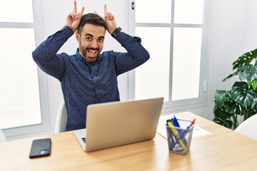 Young hispanic man with beard working at the office with laptop posing funny and crazy with fingers on head as bunny ears, smiling cheerful