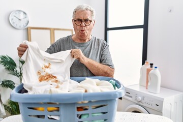 Senior caucasian man holding dirty t shirt with stain making fish face with mouth and squinting eyes, crazy and comical.