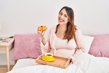 Young pregnant woman having breakfast sitting on bed at bedroom