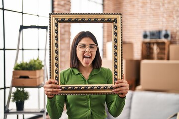 Young hispanic woman at new home holding empty frame sticking tongue out happy with funny...
