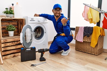 Young indian technician working on washing machine smiling with happy face looking and pointing to...