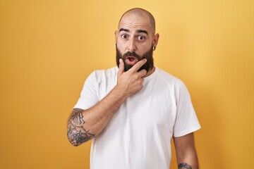 Young hispanic man with beard and tattoos standing over yellow background looking fascinated with disbelief, surprise and amazed expression with hands on chin
