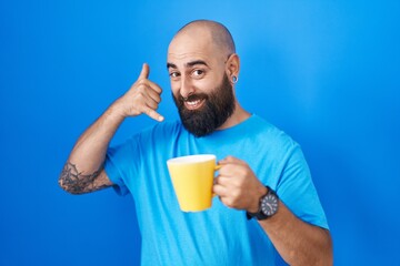 Young hispanic man with beard and tattoos drinking a cup of coffee smiling doing phone gesture with hand and fingers like talking on the telephone. communicating concepts.