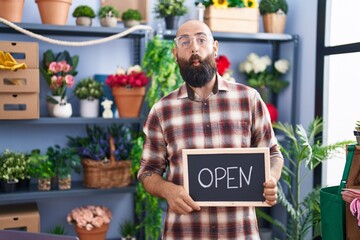 Young hispanic man with beard and tattoos working at florist holding open sign making fish face with mouth and squinting eyes, crazy and comical.