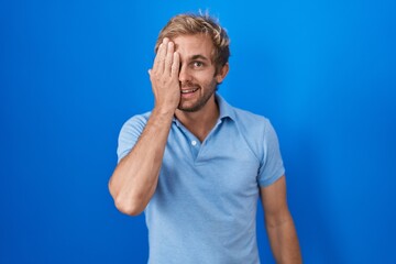 Caucasian man standing over blue background covering one eye with hand, confident smile on face and surprise emotion.
