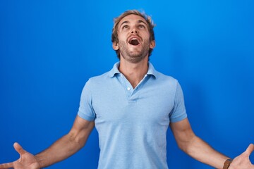 Caucasian man standing over blue background crazy and mad shouting and yelling with aggressive expression and arms raised. frustration concept.