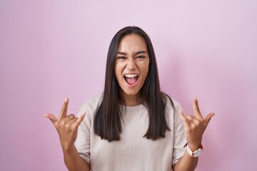 Young hispanic woman standing over pink background shouting with crazy expression doing rock symbol with hands up. music star. heavy music concept.