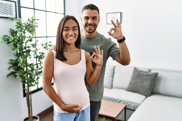 Young interracial couple expecting a baby, touching pregnant belly smiling positive doing ok sign with hand and fingers. successful expression. - obrazy, fototapety, plakaty