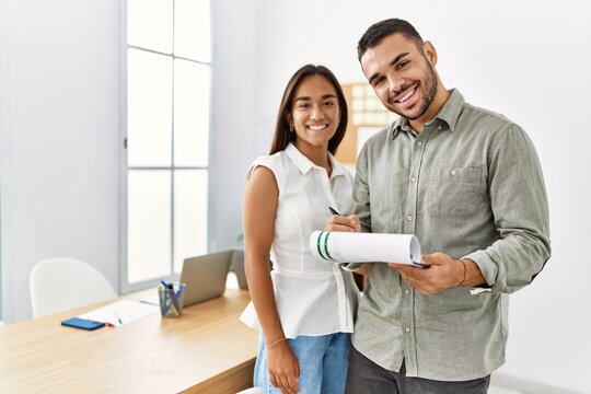 Two latin business workers smiling happy writng on clipboard working at the office.