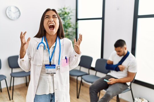 Young asian doctor woman at waiting room with a man with a broken arm crazy and mad shouting and yelling with aggressive expression and arms raised. frustration concept.