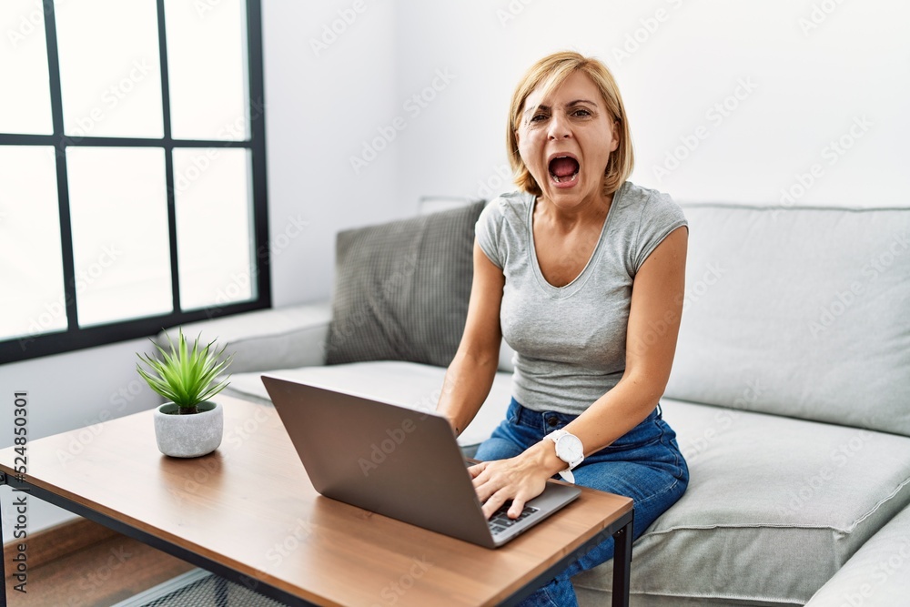 Sticker Middle age blonde woman using laptop at home angry and mad screaming frustrated and furious, shouting with anger. rage and aggressive concept.